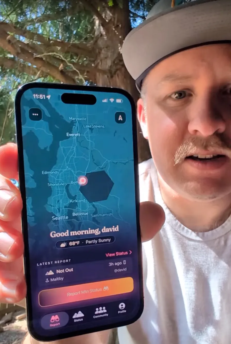 David holding up his phone outdoors under a tree, showing the Is The Mountain Out app's map screen: a “Good morning, david” greeting, his @david handle, a “Not Out” report at Maltby, and the local weather.
